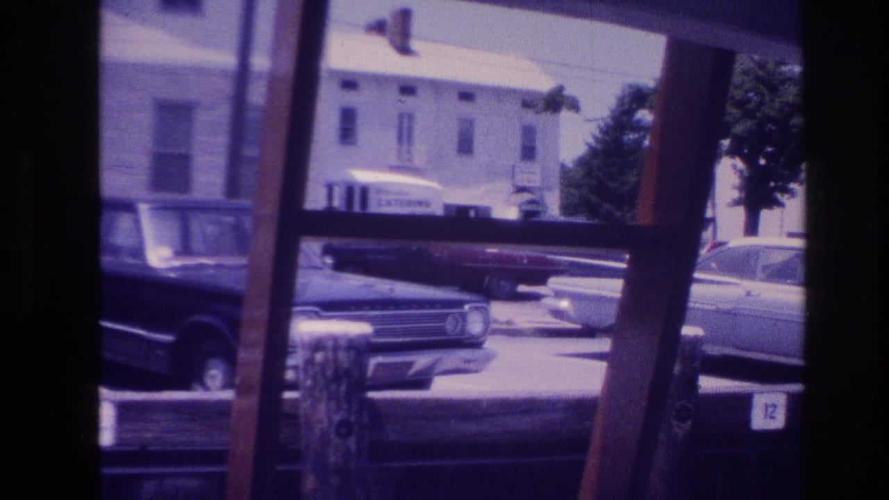 1968 Ocean City home-movie frame showing parked classic cars through a wooden frame, with a building and catering truck across the street.