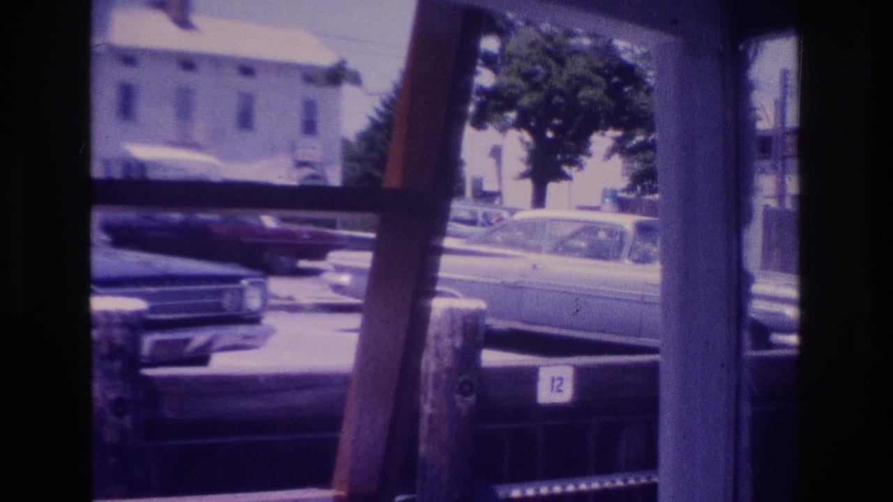 Parked cars and a street are viewed through dark vertical framing and window-like openings.