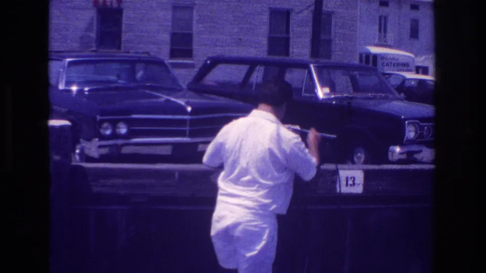 1968 Ocean City home-movie frame of a man arranging equipment near classic cars, with a building and catering truck behind him.