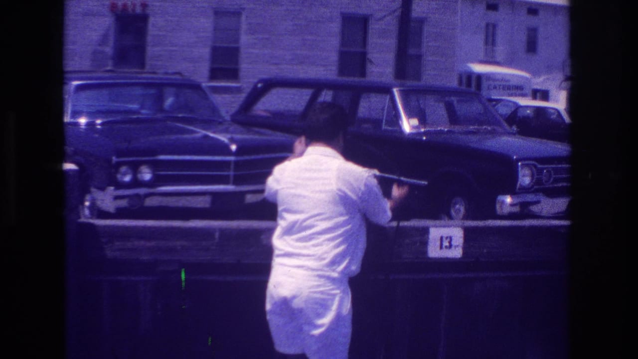 In 1968 Ocean City, New Jersey footage, a man in a white shirt and shorts stands near a wooden pier rail with classic cars parked behind.
