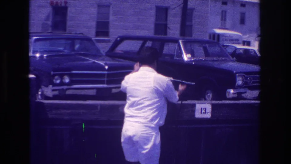 In 1968 Ocean City, New Jersey footage, a man in a white shirt and shorts stands near a wooden pier rail with classic cars parked behind.
