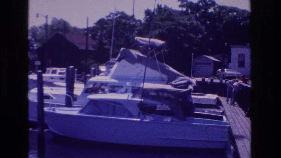 1968 Ocean City, New Jersey marina view with a moored cabin boat, wooden dock, vertical posts, trees, and shore buildings in soft 8mm color.