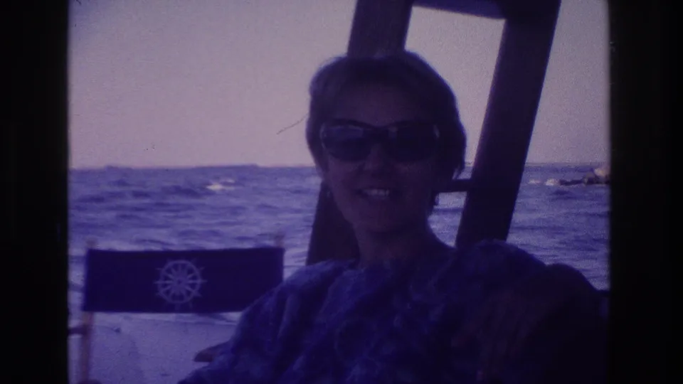 In the 1968 Ocean City boat scene, a woman with short hair and sunglasses smiles toward the camera beside a director-style chair with a ship's wheel emblem.