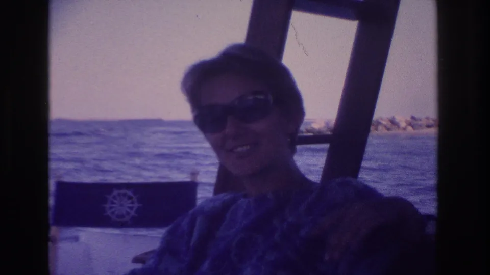 Home-movie frame of the woman turned slightly in the boat seat, wearing sunglasses against a coastal water background.