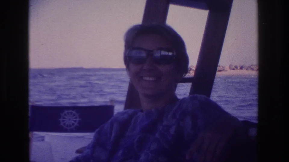 1968 seaside boating frame showing a smiling woman in sunglasses, with water and distant rocks beyond the boat.