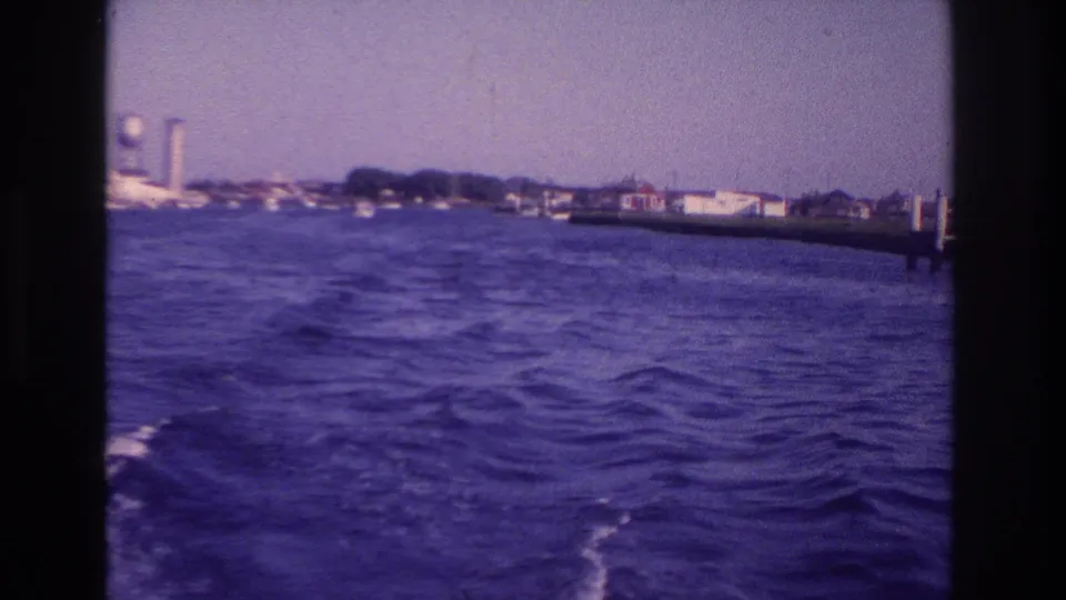 Moving-boat view over rough blue water toward the Ocean City shoreline, with waterfront buildings and a tall water tower under a hazy sky.