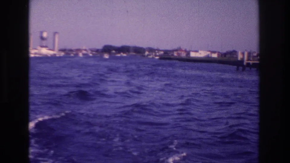 A soft 8mm frame shows a boat passenger's view across choppy water toward Ocean City shoreline structures and a water tower.
