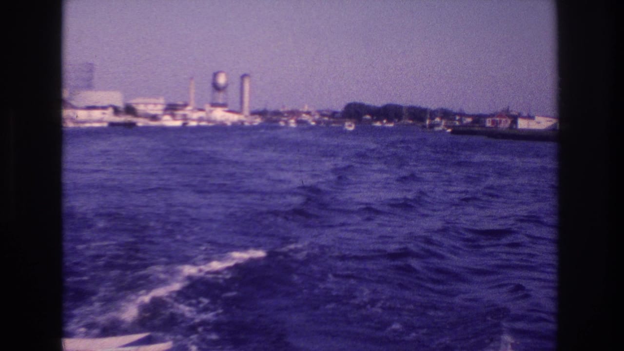 Choppy water and boat wake facing a distant shoreline with a water tower and industrial structures.
