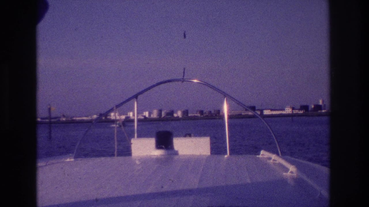 1968 Ocean City, New Jersey frame from the bow of a motorboat, with a curved rail, bay water, and distant shoreline buildings.