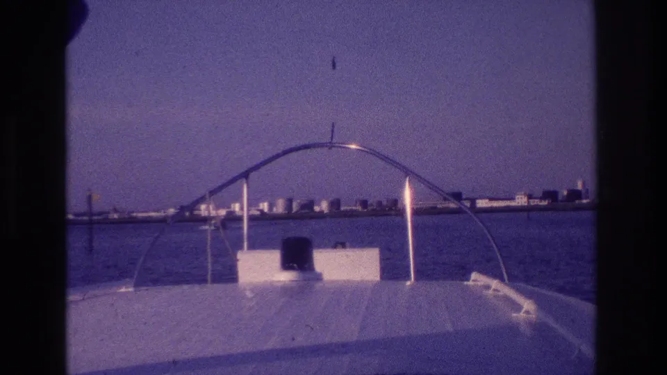 1968 Ocean City, New Jersey frame from the bow of a motorboat, with a curved rail, bay water, and distant shoreline buildings.