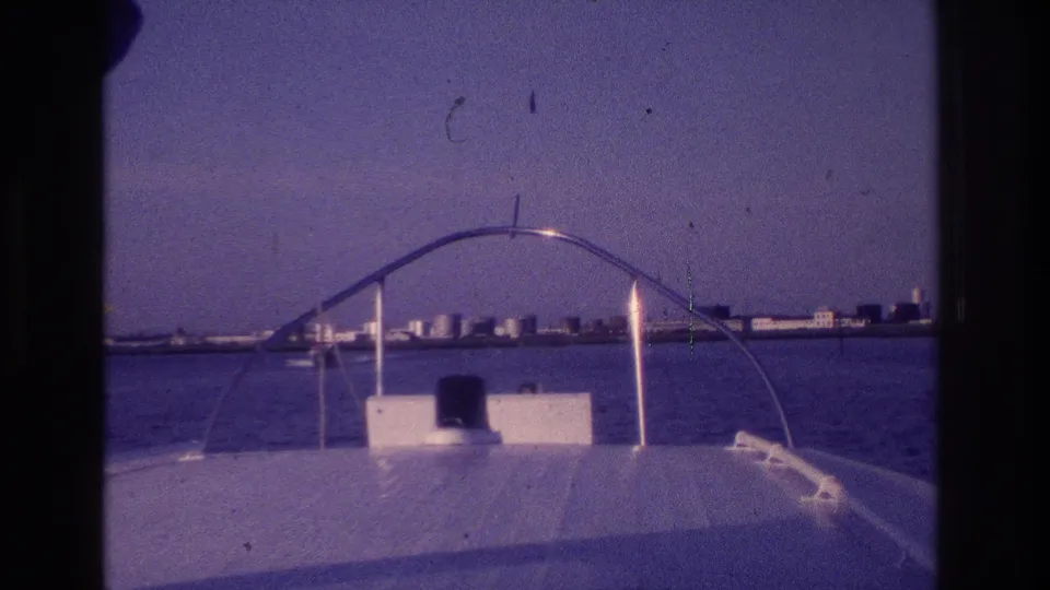 Aged 8mm frame from a motorboat in Ocean City, New Jersey, showing the bow rail against bay water and distant shoreline buildings.