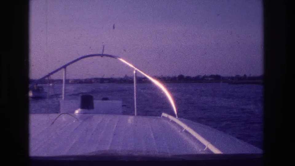 1968 home-movie frame looking over a small motorboat bow on calm Ocean City water, with shoreline and sky beyond.