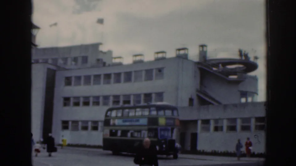 1961 Dublin Airport exterior with a double-decker bus, white terminal facade, pedestrians, and overcast sky.