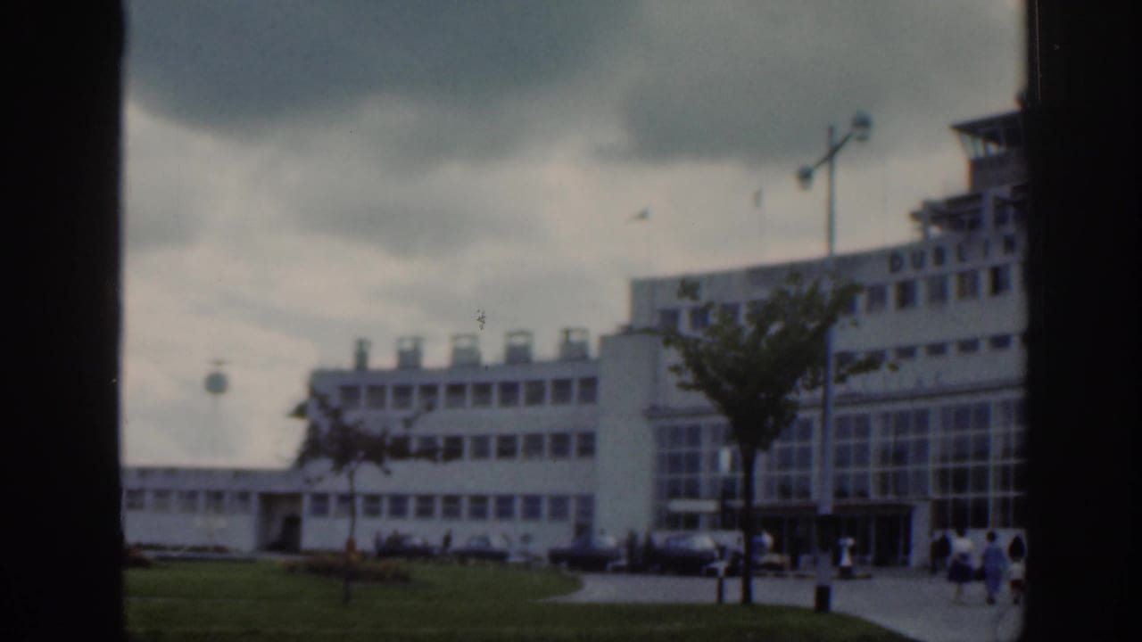 Dublin Airport terminal exterior in 1961 with modernist buildings, lawn, walkway, lamp posts, and small groups of people.