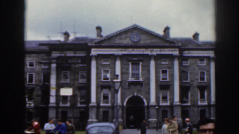 1961 Dublin footage frame showing the front of a formal stone building, with people walking and standing near the entrance.