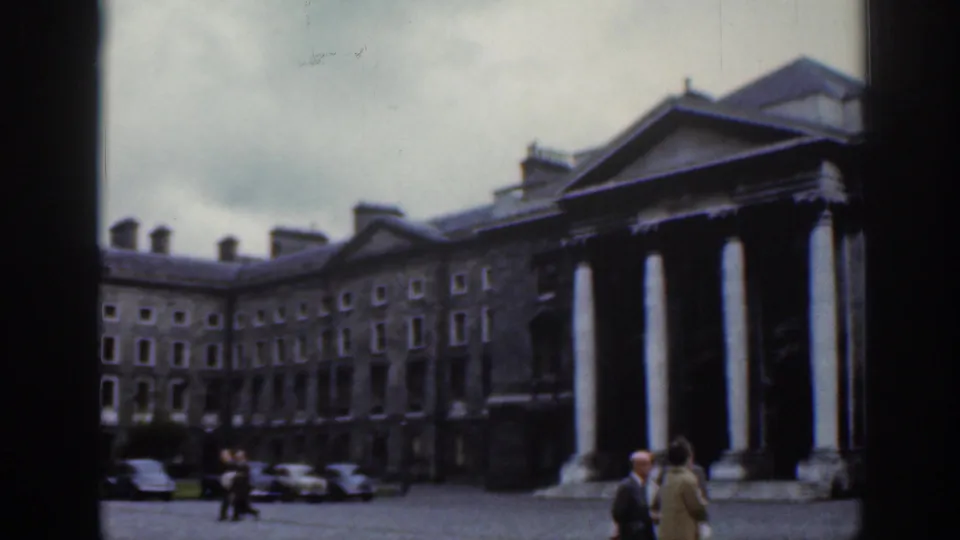 Dublin 1961 home-movie frame showing a courtyard bordered by stone buildings and a columned portico, with pedestrians and parked cars.