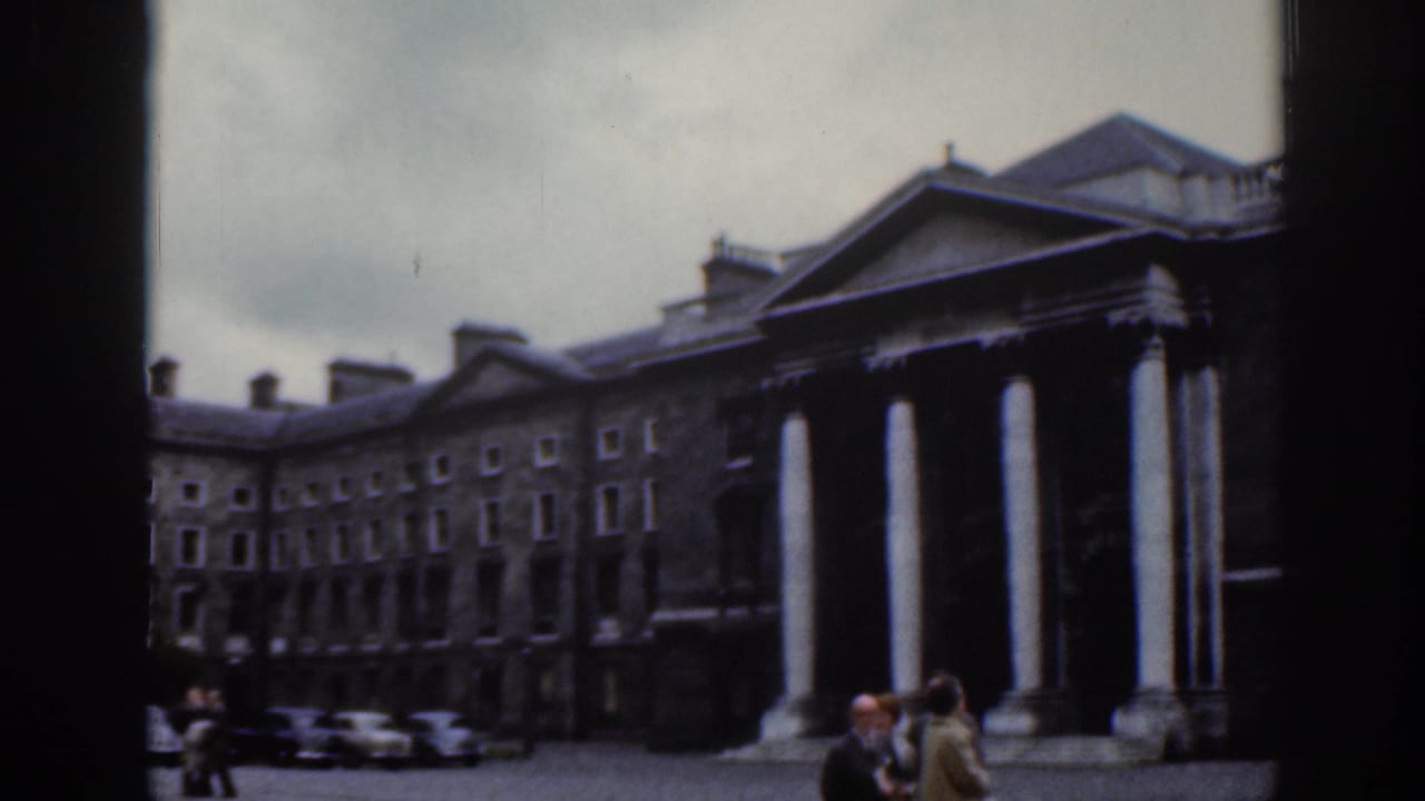 Columned stone building and courtyard with pedestrians under a cloudy sky.
