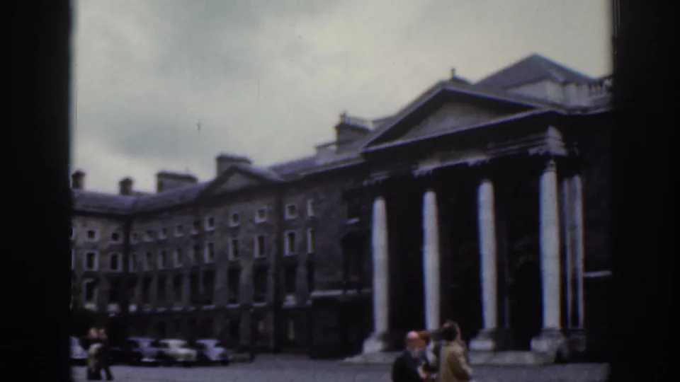 1961 Dublin home-movie frame showing a grand stone building with white columns, courtyard visitors, parked cars, and overcast sky.