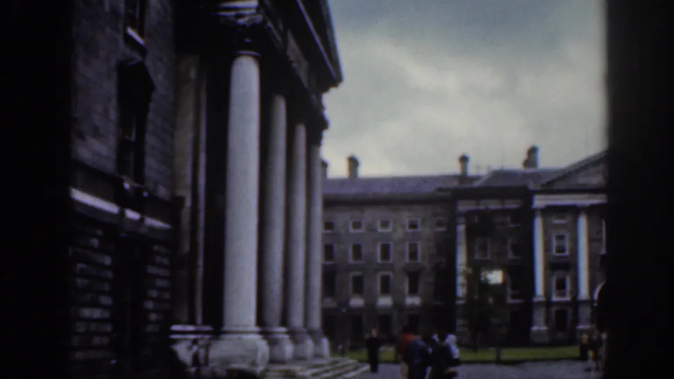 1961 Dublin frame showing a columned stone facade, courtyard lawn, pedestrians, and a larger stone building in the background.