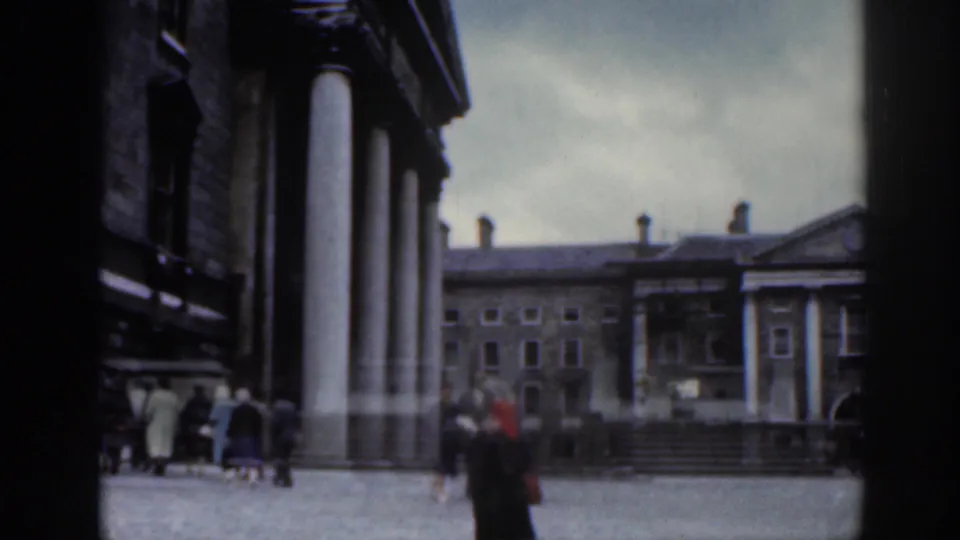 1961 Dublin home-movie view of pedestrians in coats crossing a cobbled public courtyard framed by stone buildings and columns.