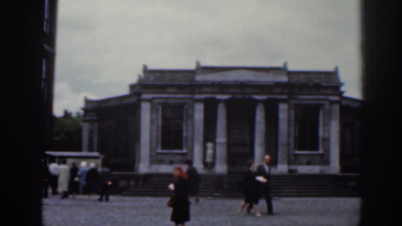 Columned stone building faces a courtyard with several pedestrians nearby.