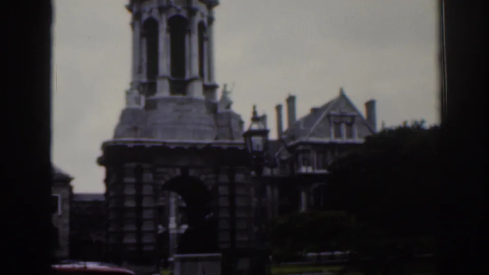 1961 Dublin home-movie frame showing a stone tower with arched openings beside gabled buildings and a street lamp.