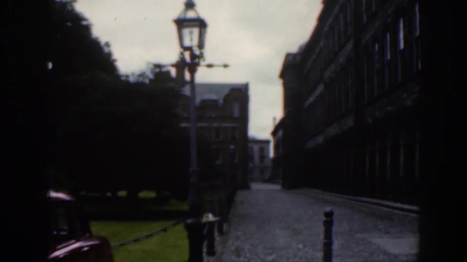 1961 Dublin street view with a tall lamppost, cobbled lane, dark stone buildings, trees, lawn, and a red car partly visible at left.
