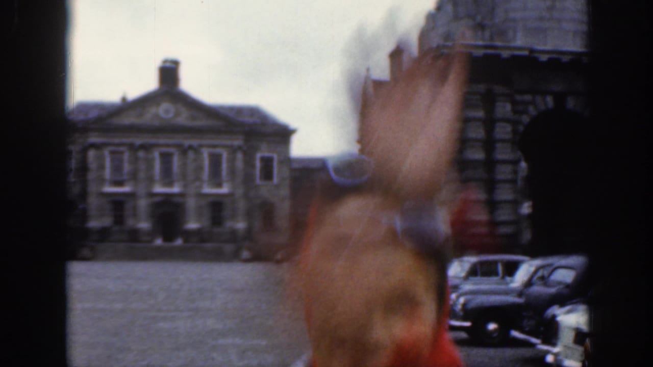 A blurred person waves close to the camera in a Dublin courtyard scene, with parked vintage cars and a classical stone building beyond.