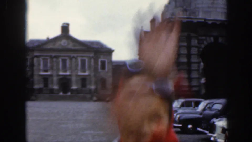 A blurred person waves close to the camera in a Dublin courtyard scene, with parked vintage cars and a classical stone building beyond.