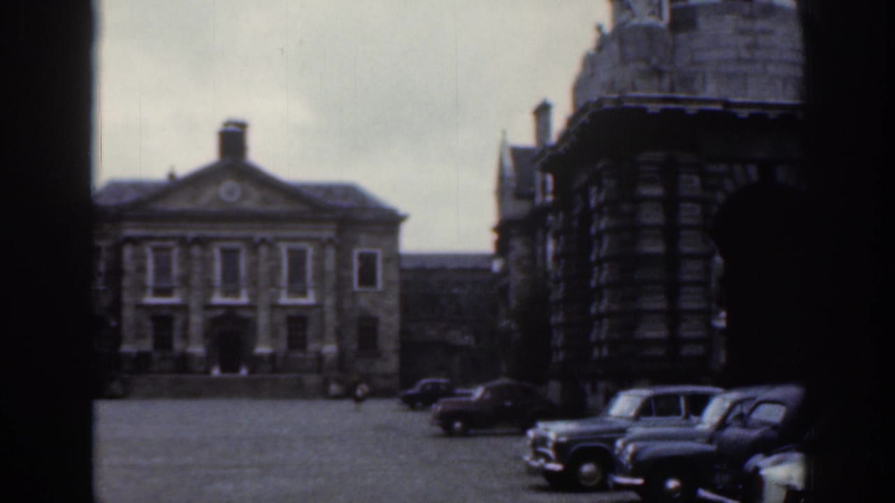 1961 Dublin home-movie frame showing a cobblestone square, stone civic buildings, and parked cars along the street.