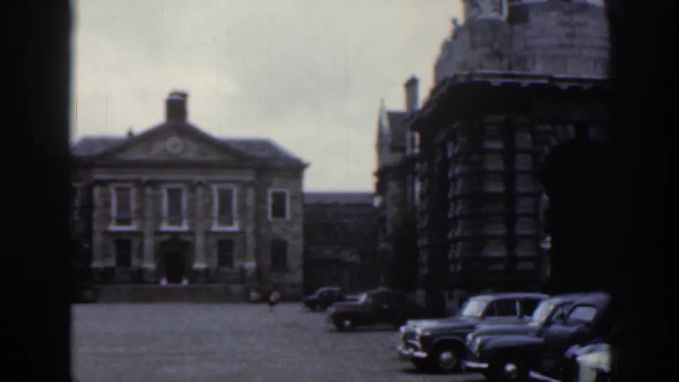 1961 Dublin home-movie frame showing a cobblestone square, stone civic buildings, and parked cars along the street.