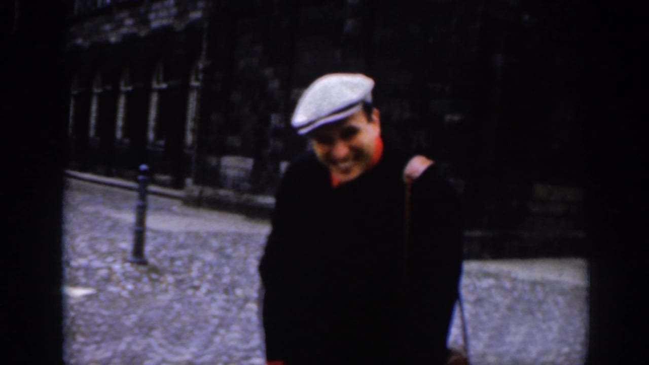 1961 Dublin street view with a smiling man in a pale flat cap and dark coat walking past stone buildings.