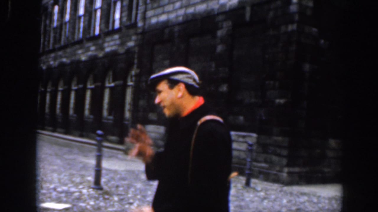 A 1961 Dublin frame shows the man in profile, gesturing while walking beside a dark stone building on a cobblestone street.
