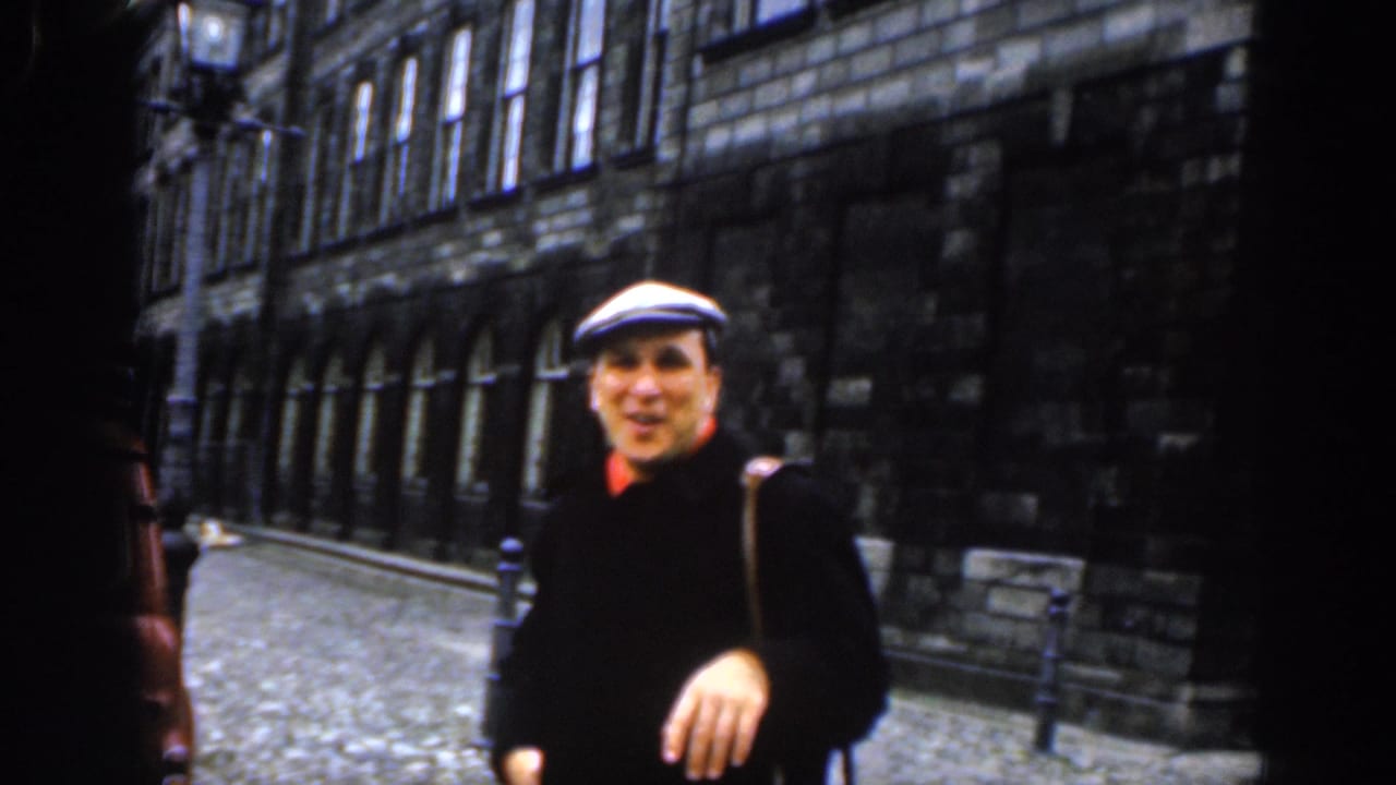 In 1961 Dublin, a man in a white flat cap, dark coat, and red collar smiles near the camera on a cobblestone street lined with stone buildings.