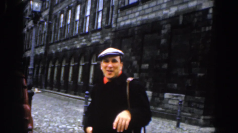 In 1961 Dublin, a man in a white flat cap, dark coat, and red collar smiles near the camera on a cobblestone street lined with stone buildings.