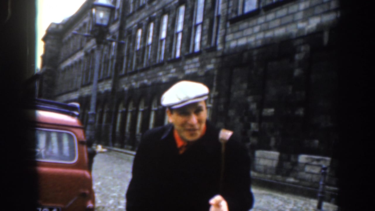 Man in a white flat cap gestures near a red vehicle on a narrow stone-lined street.