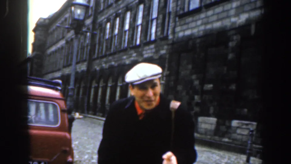The man in the white flat cap appears to gesture as he stands near a red vehicle on a Dublin street with dark stone buildings and arched windows.