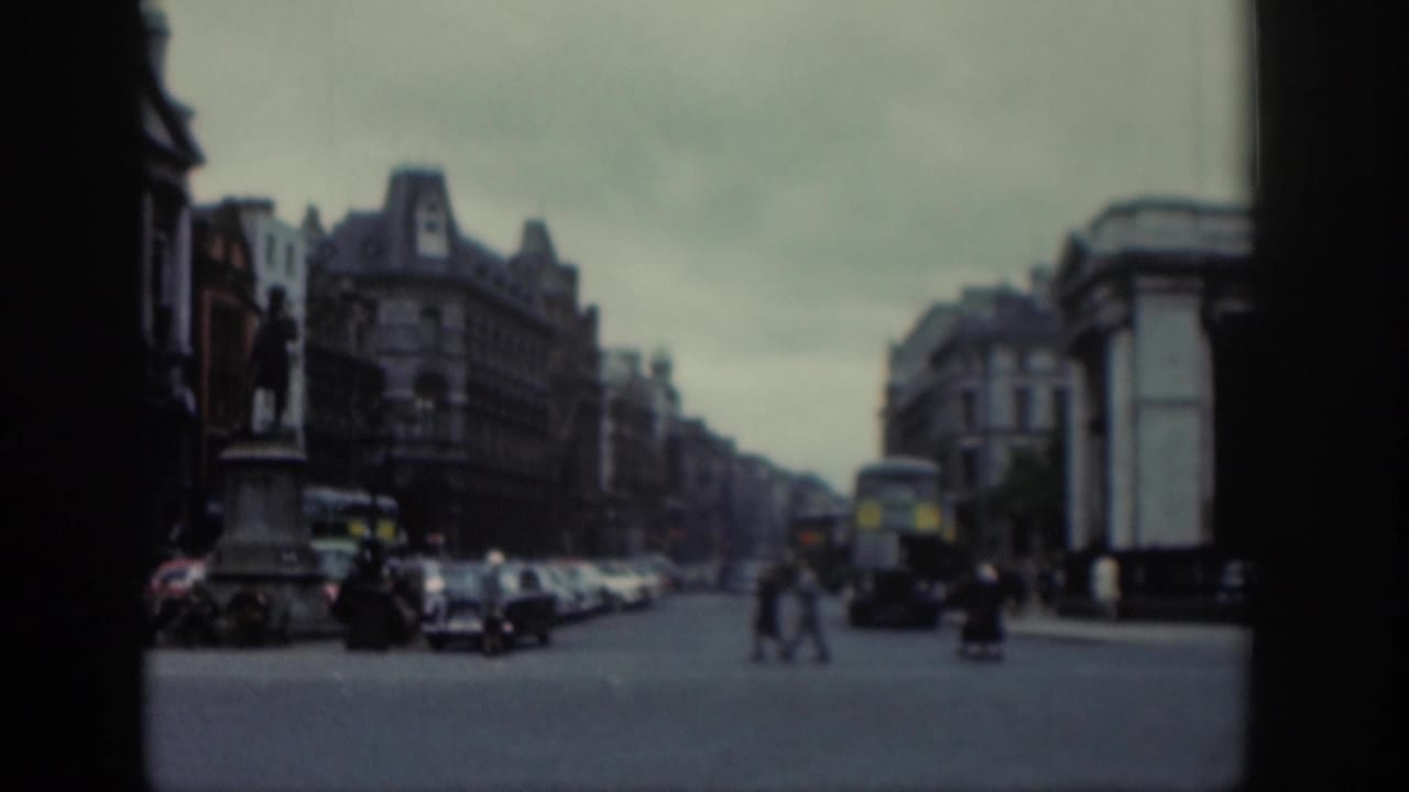 1961 Dublin home-movie view of a broad street intersection with a statue, period cars, pedestrians, and a double-decker bus.