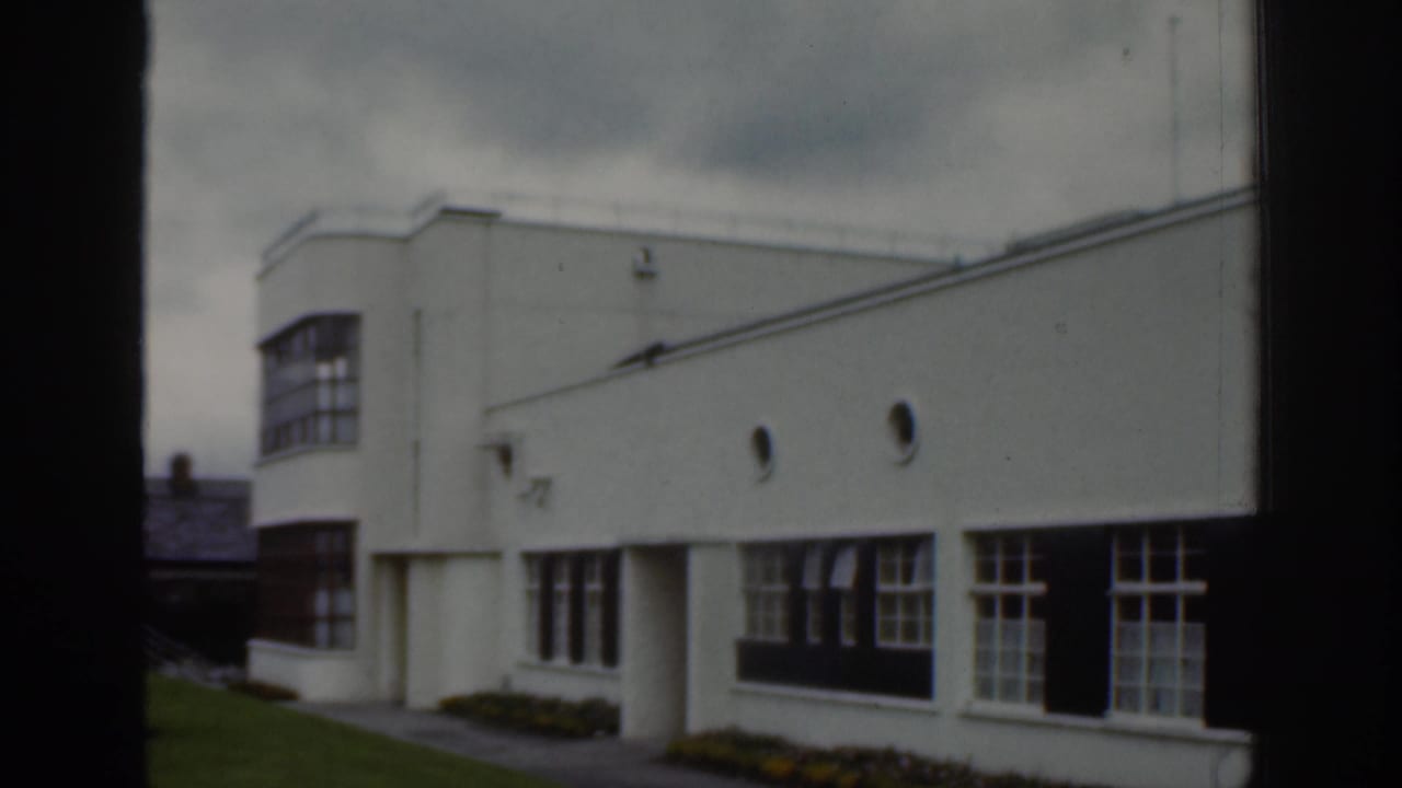 Dublin 1961 home-movie frame showing a white modernist building exterior with broad windows, lawn, and overcast sky.
