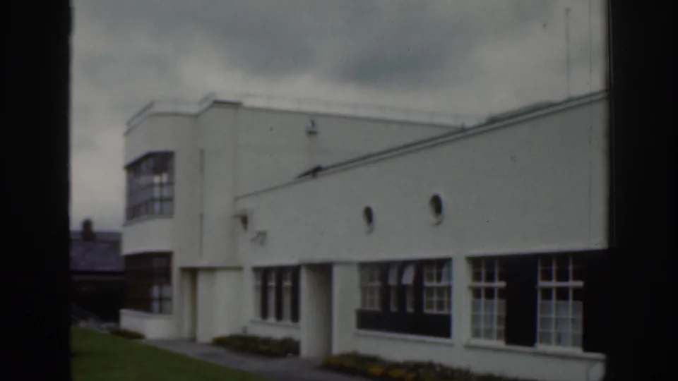 Dublin 1961 home-movie frame showing a white modernist building exterior with broad windows, lawn, and overcast sky.