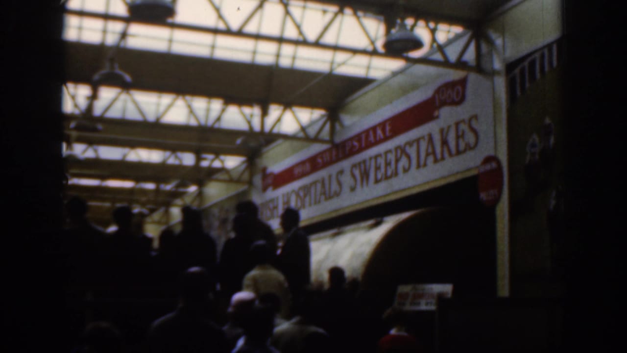Dublin 1961 interior frame with silhouetted people beneath skylights and a large Irish Hospitals Sweepstakes banner.