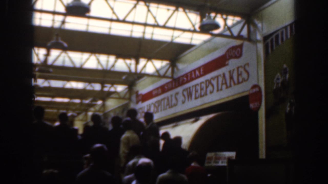 Dublin 1961 home-movie frame of people gathered in a hall under steel trusses and the Irish Hospitals Sweepstakes sign.