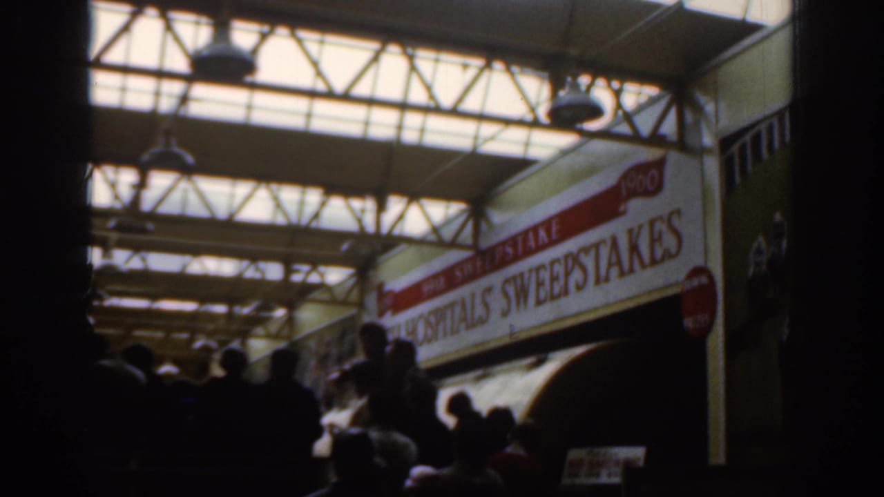1961 Dublin home-movie frame showing pedestrians under a covered walkway, a large sweepstakes sign, and city traffic outside.