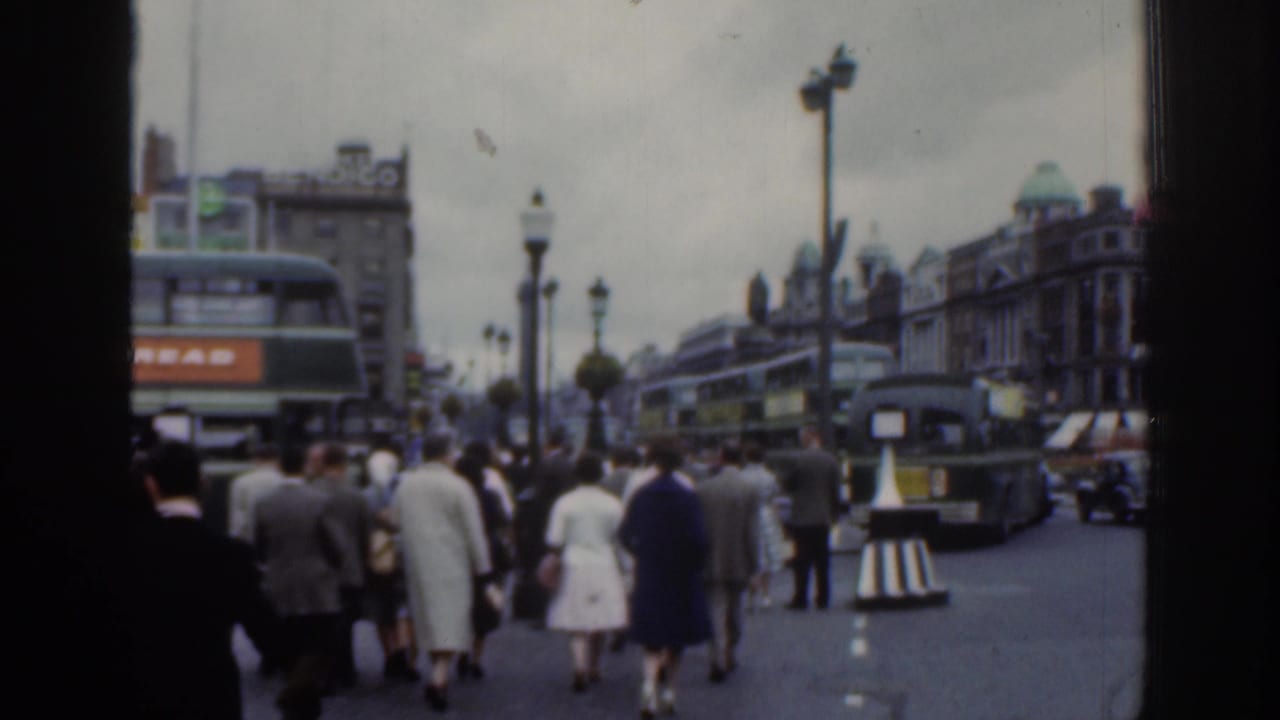 Crowded street with pedestrians, double-decker buses, cars, lamps, and older buildings.