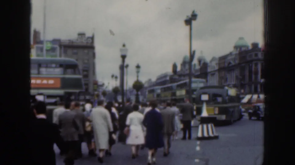 1961 Dublin street view with pedestrians walking among double-decker buses, cars, street lamps, and historic-looking commercial buildings.