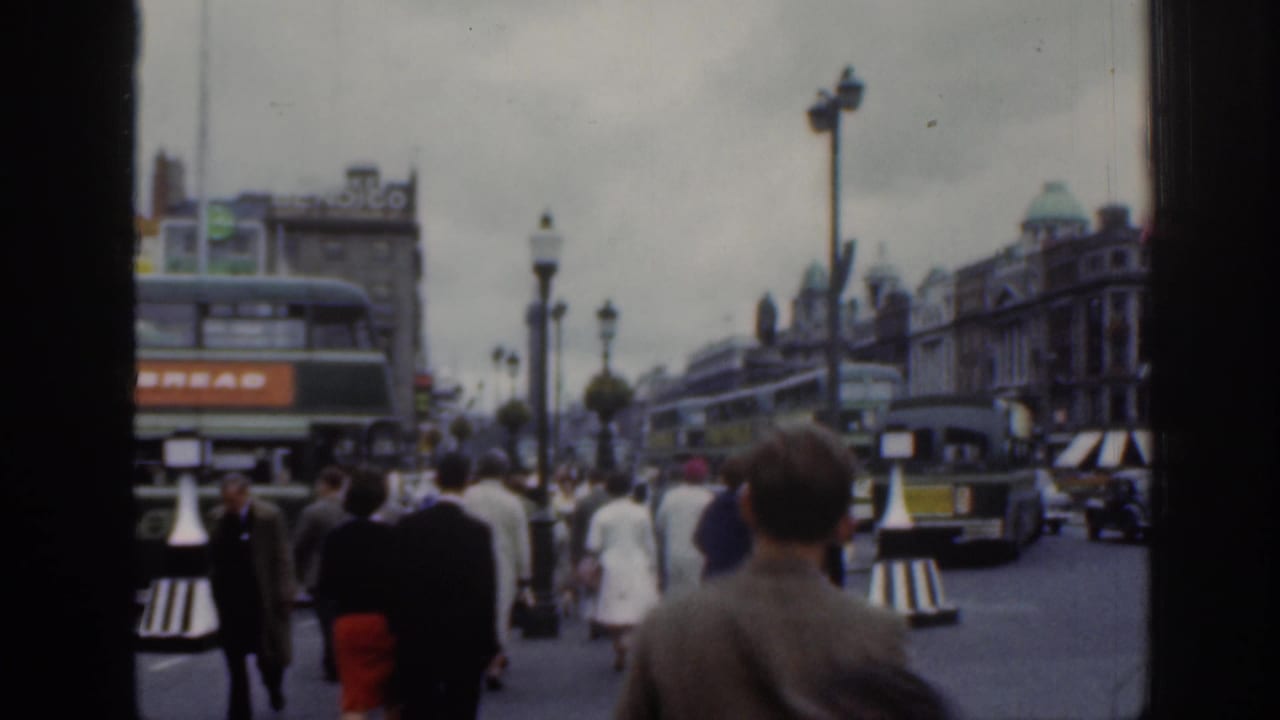 1961 Dublin home-movie street frame looking past pedestrians toward double-decker buses, cars, lamps, and dense city buildings.