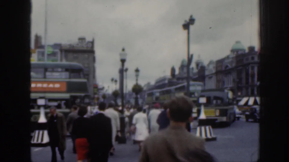 1961 Dublin home-movie street frame looking past pedestrians toward double-decker buses, cars, lamps, and dense city buildings.