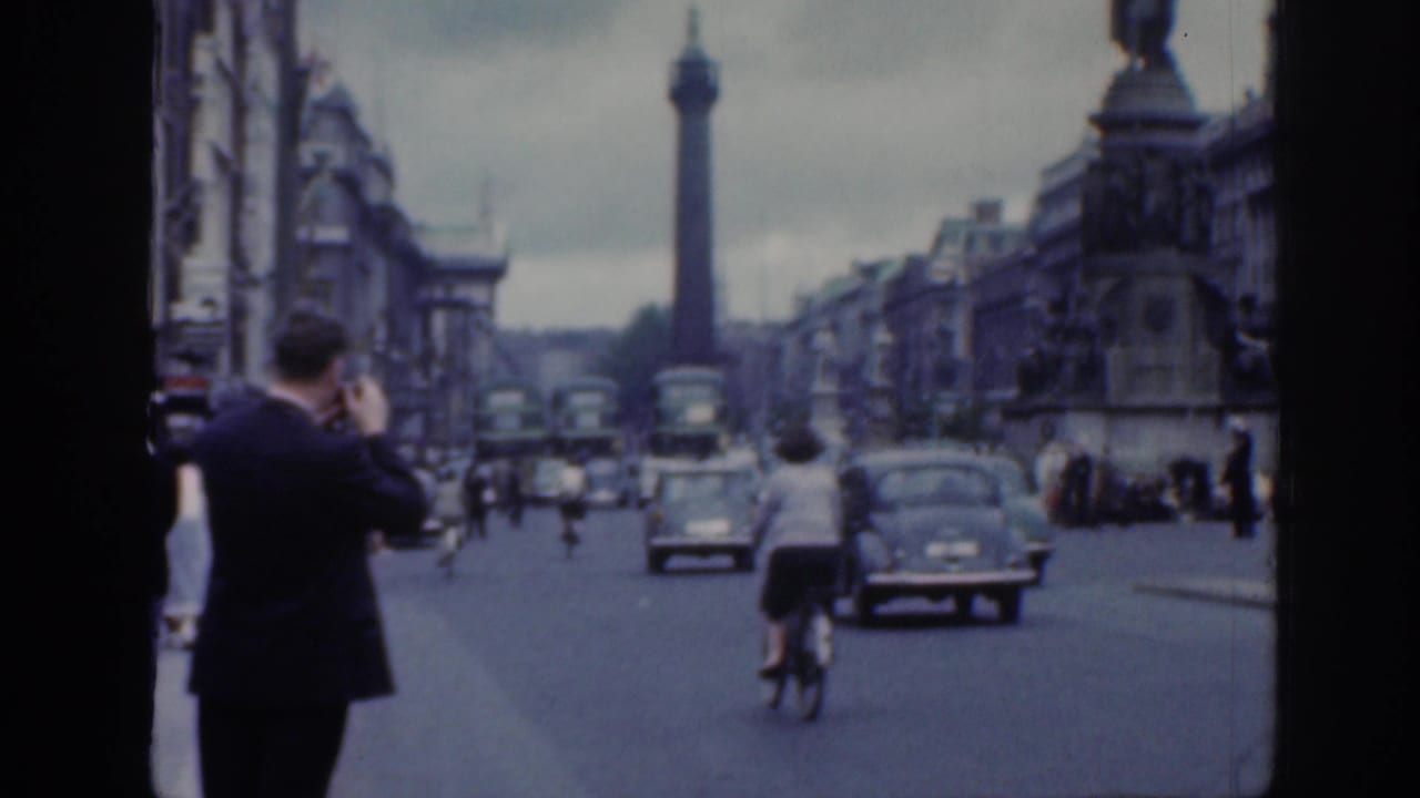 1961 Dublin street scene with a tall column monument in the distance, a cyclist, vintage cars, buses, pedestrians, and ornate buildings.
