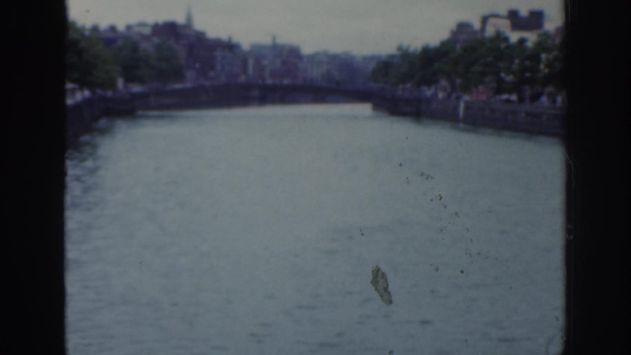 1961 Dublin home-movie frame showing a river from a bridge, with stone embankments, trees, distant buildings, and overcast light.