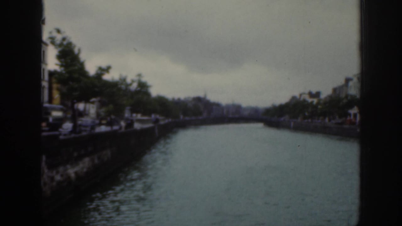 1961 Dublin home-movie view along the river, showing a stone quay, roadside vehicles, trees, and distant riverside buildings.